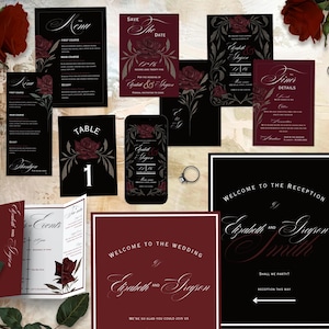 May include: A selection of wedding stationery in black and claret, with rose floral designs. Includes menu cards, save-the-date cards, and reception details. A smartphone displays a matching invitation design. A ring is also present.
