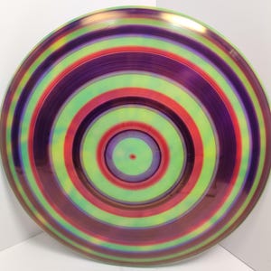 May include: A circular disc with a multi-coloured concentric circle pattern. The colours are shades of green, red, purple, and yellow. The centre of the disc is a smaller circle with a green centre and purple outer ring.