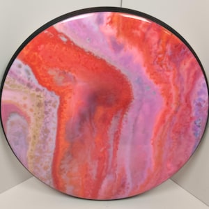 May include: Round abstract art painting with a black frame. The painting features a swirling pattern of pink, red, and white colors.