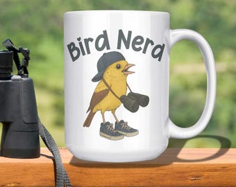 May include: A white ceramic mug with the words "Bird Nerd" and a cartoon bird wearing a hat, sneakers, and binoculars. Black binoculars with a neck strap are next to the mug. The items are on a wooden surface.