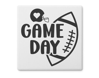 May include: Square white ceramic tile with a black graphic. The graphic features a football helmet with a heart, a football, and the words "GAME DAY" in a bold, sans-serif font. The design is simple and modern.