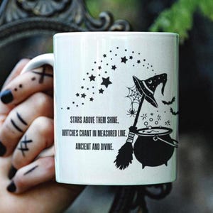 May include: White ceramic mug with a black witch-themed design. The design includes a witch's hat, broom, cauldron, bats, and stars. Text on the mug reads: "STARS ABOVE THEM SHINE, WITCHES CHANT IN MEASURED LINE, ANCIENT AND DIVINE."