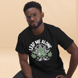 May include: Black t-shirt with a graphic of a cannabis leaf character wearing headphones and the text "LEAF ME ALONE" in white. The character has an angry expression and is surrounded by cannabis leaves.
