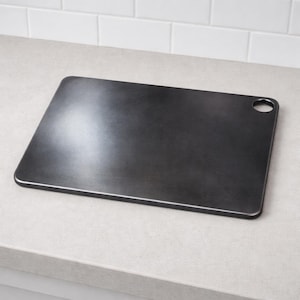 May include: A rectangular black cutting board with rounded corners and a circular hole in the top right corner for hanging. The surface has a smooth, matte finish with subtle light reflections across the center of the board.