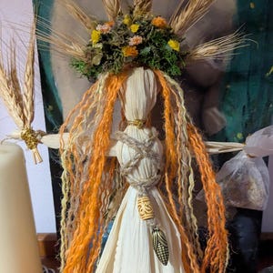 May include: A handcrafted corn husk doll with long orange and yellow yarn hair, wearing a white dress. The doll has a wheat crown and is adorned with a decorative pendant. The doll is part of a spiritual or folk art theme.