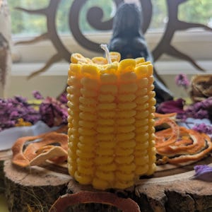 May include: A yellow, textured candle shaped like a corn cob, with a white wick. The candle sits on a wooden slice, surrounded by dried orange peel and purple flowers. A decorative metal sun and a black statue are in the background.