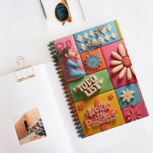 Puede incluir: Un cuaderno de espiral colorido con un diseño caprichoso. La portada presenta una cuadrícula de cuadrados con adornos en 3D, incluyendo una niña, flores y las palabras "Ideas", "To Do List" y "Work in Progress". El cuaderno está abierto sobre una superficie blanca.