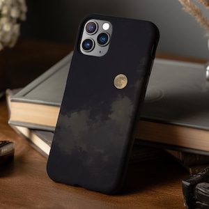 Puede incluir: Funda de teléfono negra con un diseño de cielo nocturno con luna llena y nubes. La funda está sobre una superficie de madera junto a una pila de libros y un pequeño objeto decorativo. La funda del teléfono tiene un acabado mate.