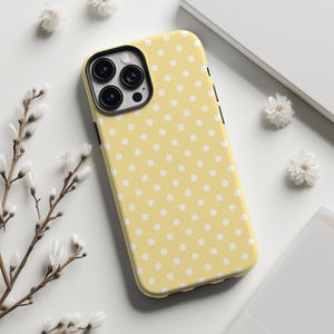 May include: A yellow phone case with a white polka dot pattern. The case has a black camera lens cutout and black side buttons. The case is surrounded by white flowers and a white background.