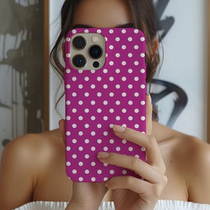 May include: A magenta phone case with a white polka dot pattern. The case has a camera cutout and is held up in front of a person. The phone case is a vibrant accessory.