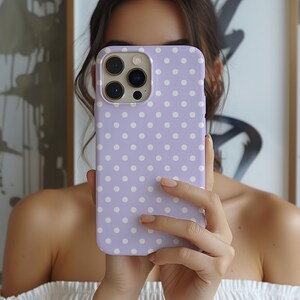 May include: A lilac phone case adorned with a white polka dot design. The case, designed for a smartphone, has a camera aperture. The case is held up in front of a person.
