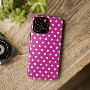 May include: A magenta phone case with a white polka dot pattern. The case has a glossy finish and a camera cutout. The phone case is on a wooden surface.