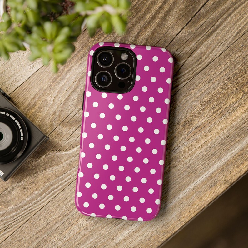 May include: A magenta phone case with a white polka dot pattern. The case has a glossy finish and a camera cutout. The phone case is on a wooden surface.