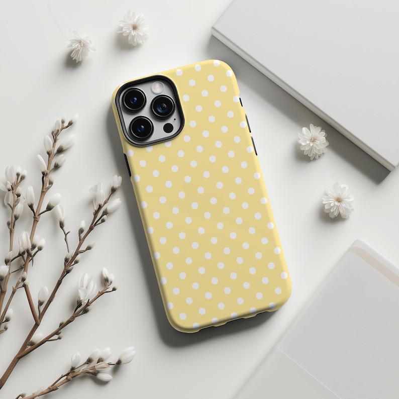 May include: A yellow phone case with a white polka dot pattern. The case has a black camera lens cutout and black side buttons. The case is surrounded by white flowers and a white background.