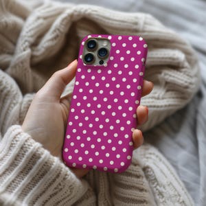May include: A magenta phone case with a white polka dot pattern. The case is held in a hand and is set against a neutral background. The phone case has a camera module with three lenses.
