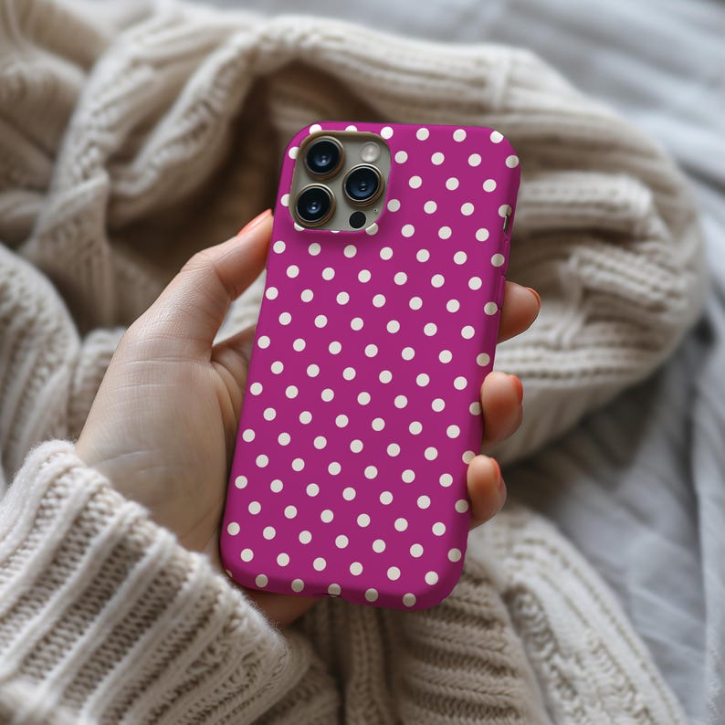May include: A magenta phone case with a white polka dot pattern. The case is held in a hand and is set against a neutral background. The phone case has a camera module with three lenses.