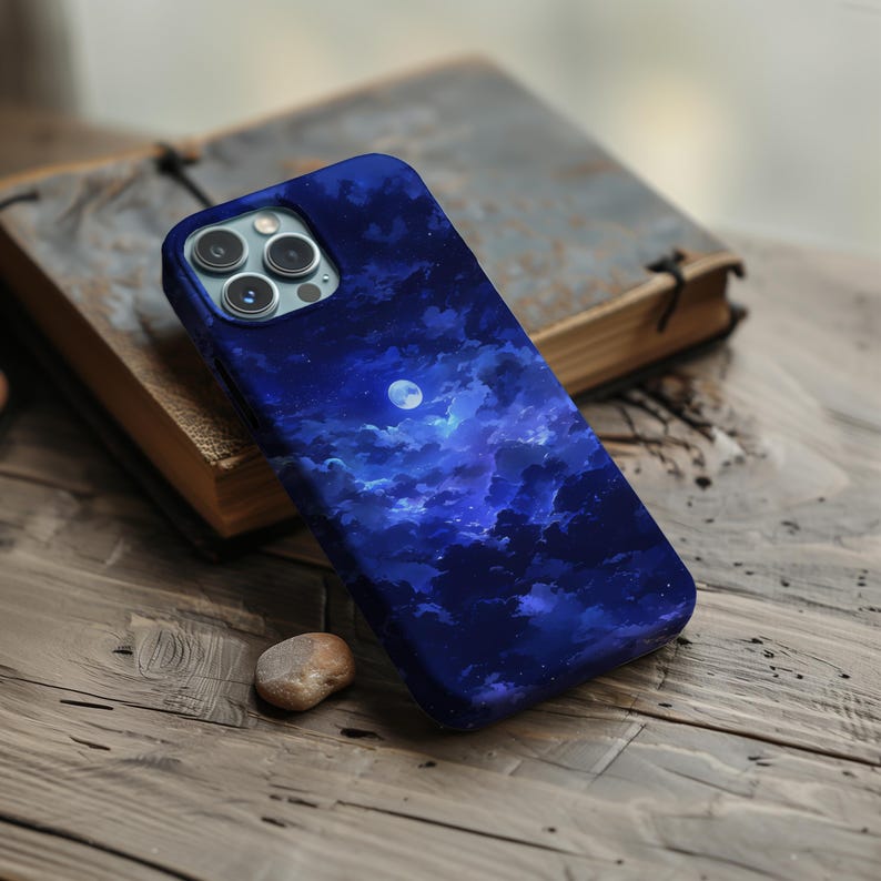 Puede incluir: Una funda de tel&eacute;fono azul oscuro con un dise&ntilde;o de cielo nocturno, con una luna llena y nubes. La funda est&aacute; sobre una superficie de madera junto a un libro. La funda tiene un m&oacute;dulo de c&aacute;mara con tres lentes.
