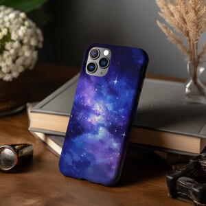 May include: A phone case with a galaxy design. The case is dark blue with purple and white clouds and stars. The case has a cutout for the camera lens.