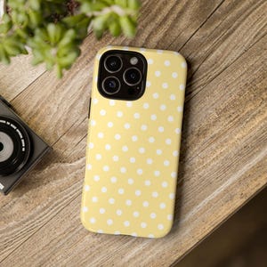 Może przedstawiać: Ż&oacute;łte etui na telefon z białym wzorem w kropki. Etui leży na drewnianej powierzchni, z aparatem i rośliną w tle. Etui ma błyszczące wykończenie i ochronną konstrukcję.