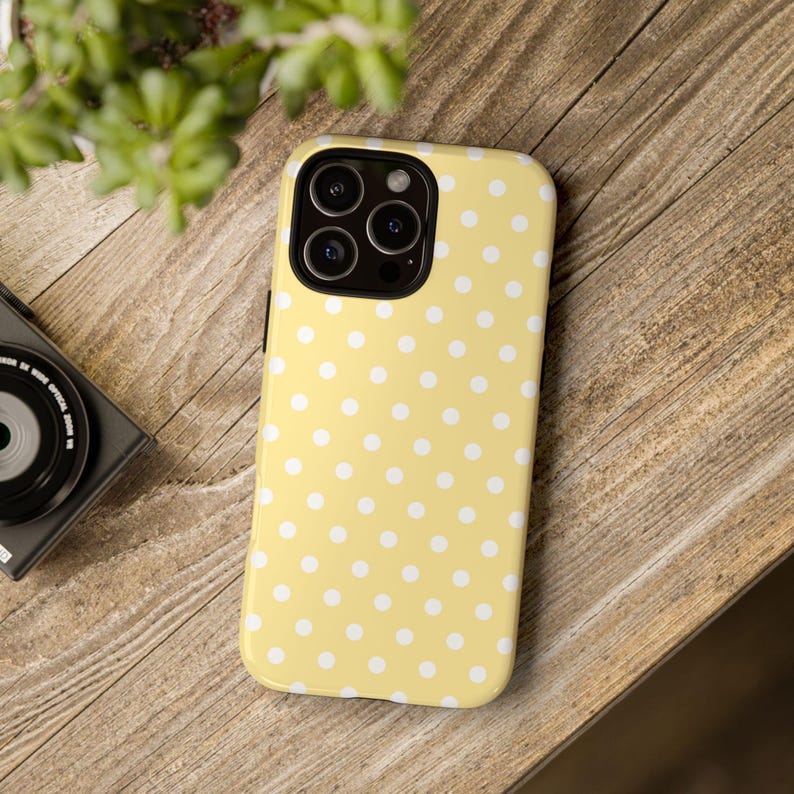 Może przedstawiać: Ż&oacute;łte etui na telefon z białym wzorem w kropki. Etui leży na drewnianej powierzchni, z aparatem i rośliną w tle. Etui ma błyszczące wykończenie i ochronną konstrukcję.