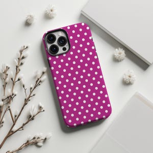 May include: A magenta phone case with a white polka dot pattern. The case has a black camera module and is surrounded by white flowers and a white background.