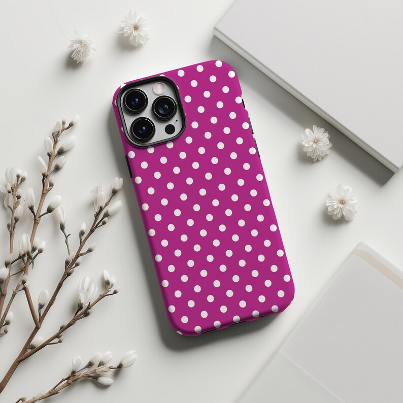 May include: A magenta phone case with a white polka dot pattern. The case has a black camera module and is surrounded by white flowers and a white background.