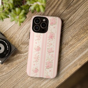 Może przedstawiać: Etui na telefon z r&oacute;żowo-białym wzorem w paski, ozdobione pionowymi rzędami r&oacute;żowych r&oacute;ż. Etui leży na drewnianej powierzchni, z aparatem i zielenią w tle.