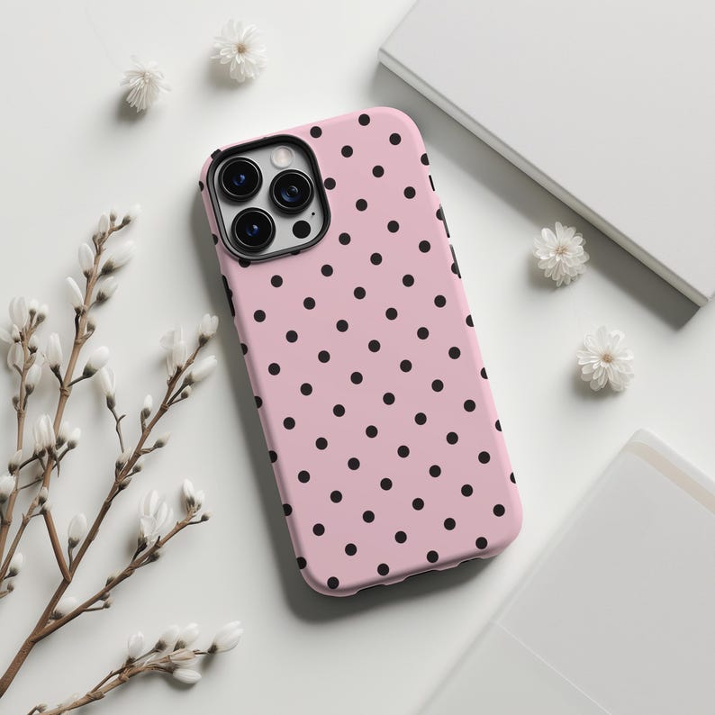 May include: A pink phone case with a black polka dot pattern. The case has a black camera lens cutout and is surrounded by white flowers and a white background.