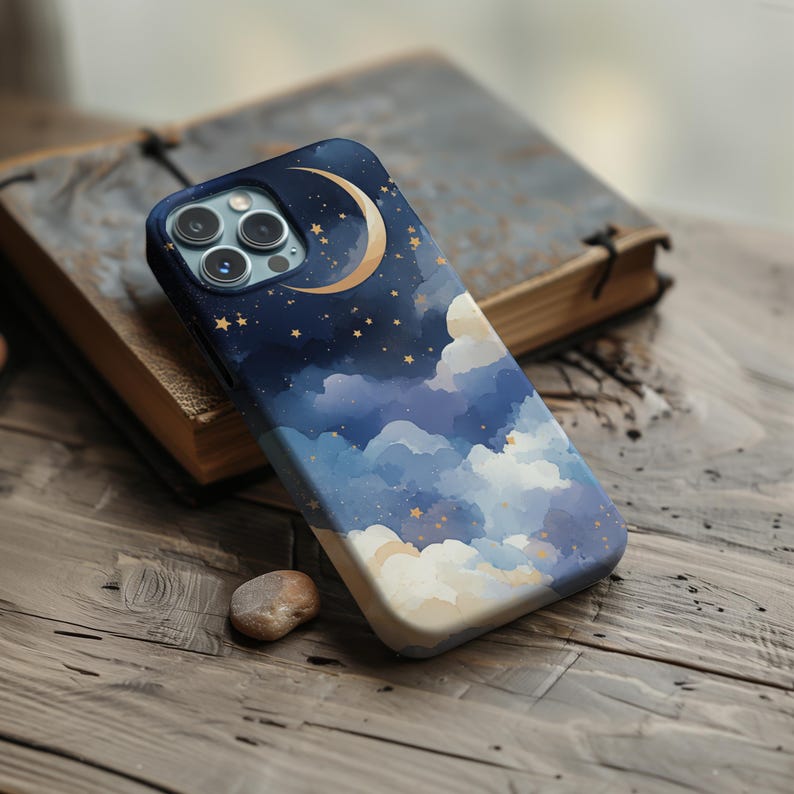 May include: A smartphone case with a celestial design featuring a crescent moon, stars, and clouds in shades of blue, white, and gold. The case is resting on a wooden surface next to a book.