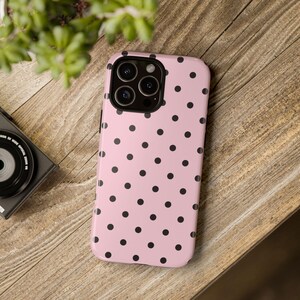 May include: A pink phone case with a black polka dot pattern. The case is on a wooden surface, with a camera and greenery in the background. The phone case has a glossy finish and a protective design.