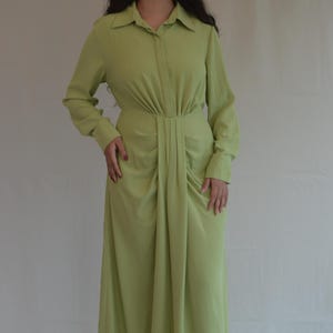 Op de afbeelding: Een lange, lichtgroene jurk met een kraag en lange mouwen. De jurk heeft een geplooid detail in de taille en een zwierige rok. Het model draagt witte sandalen. De jurk is gemaakt van een zachte, soepele stof.