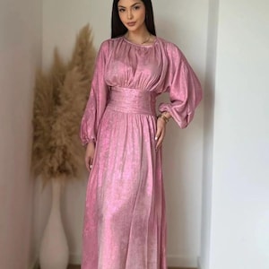 Puede incluir: Un vestido largo de satén rosa con cintura ajustada y mangas largas. El vestido tiene un brillo sutil y una falda fluida.