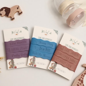 May include: Three baby name reminder cards with names in purple, blue, and brown bands. The cards have a cartoon illustration of a fox with gifts. A wooden lion and a baby bottle are also in the image.