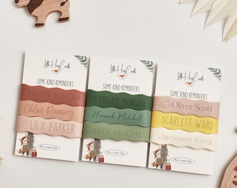 May include: Three cards with personalized name bracelets in shades of pink, green, and yellow. Each bracelet has a name engraved on it. The cards have the text "Some Kind Reminders" and a logo. A wooden lion toy is in the background.