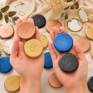 May include: A collection of round, metal compact mirrors in various colors including rose gold, gold, blue, and black. Each mirror features a single engraved script letter on the front cover. Some mirrors are closed, while one is open to reveal the glass.