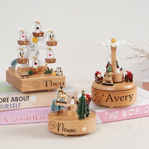May include: Wooden Christmas-themed music boxes with personalized names. One features a Ferris wheel design with snowman figures in the gondolas. Another has a carousel design with miniature trees and figures. The third has a snowman on a swing. Each box has a name engraved.