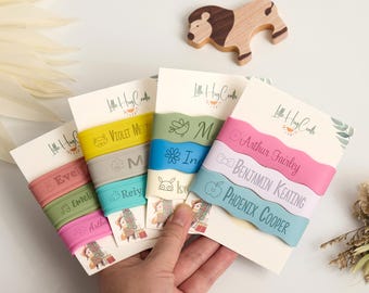 May include: Four sets of colourful silicone bracelets with names and illustrations are held in a hand. Each set is attached to a card with the text "Little Hug Crafts". A wooden lion toy is in the background.