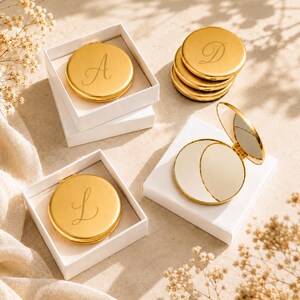 May include: Several round gold-toned compact mirrors with engraved script letters A, D, and L. Some mirrors are closed and placed inside square white gift boxes, while one mirror is open to reveal the reflective surface and gold-toned metal casing.
