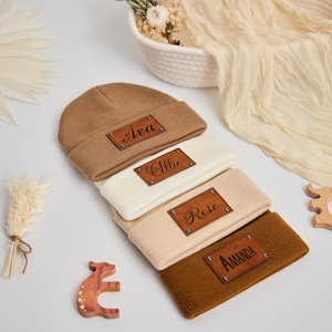 May include: Four knitted beanies in shades of brown, cream, and tan, each with a rectangular leather patch. The patches are engraved with the names "Ava", "Allie", "Rose", and "Amanda". The beanies are stacked on a white surface.