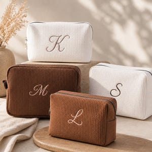 Puede incluir: Cuatro estuches rectangulares con cremallera hechos de tela con textura de gofre. Dos estuches son blancos con las letras bordadas K y S, y dos son marrones con las letras bordadas M y L. Cada estuche tiene una cremallera metálica en la parte superior.