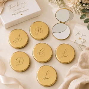 May include: Six round gold-toned metal compact mirrors, each engraved with a unique cursive letter. One mirror is open, revealing two reflective surfaces. A white gift box is present with the text Thank you FOR BEING MY BRIDESMAID.