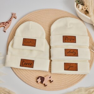 Puede incluir: Gorros de color crema con etiquetas de nombre de cuero personalizadas. Un juego incluye un gorro de bebé a juego. Los nombres "Carrie" y "Kimberley" se muestran en un juego, y "Raisin", "Ainsley" y "Zoe" en el otro.