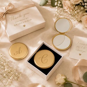 May include: A set of round gold-toned compact mirrors with engraved letters A and C. One mirror is open, revealing two reflective surfaces. The items are presented in a white gift box with the text Thank you FOR BEING MY BRIDESMAID.
