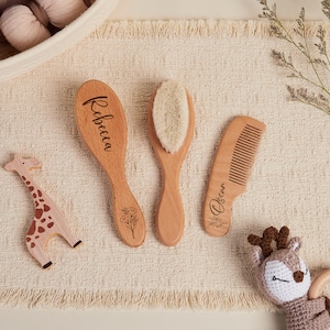 Könnte beinhalten: Ein Baby-Pflegeset aus Holz mit einer Bürste mit weichen Borsten, einem Kamm und einer Bürste mit dem Namen "Rebecca" und einem Blumendesign. Ein Holzgiraffe und ein Häkeltier-Spielzeug sind ebenfalls vorhanden.