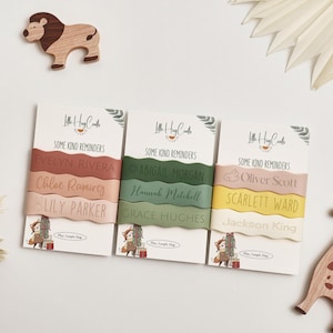 Peut inclure: Trois ensembles de bracelets personnalisés en silicone rose, vert et jaune, chacun avec un nom. Les cartes indiquent "Some Kind Reminders" et présentent une petite illustration d'un renard avec un cadeau. Un jouet en bois en forme de lion est en arrière-plan.