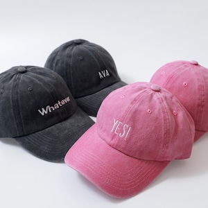 May include: Four baseball caps in shades of gray and pink. The gray caps have the words "Whatever" and "AVA" embroidered in white. The pink caps have the word "YES!" embroidered in white.