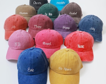 May include: A collection of colorful baseball caps in various shades, including red, blue, and yellow. Each cap features embroidered text, with names and phrases like "Owen," "Evie," and "Go Sports." The caps are arranged on a white surface.