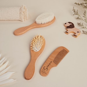 Op de afbeelding: Een houten haarverzorgingsset voor baby's met een borstel met zachte borstelharen, een kam met de naam "Sienna" en een borstel met houten pinnen. Een houten leeuwenspeelgoed en een opgerolde washand zijn ook aanwezig.