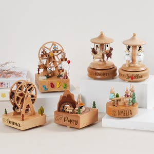May include: A collection of wooden music boxes with various themes, including a Ferris wheel, carousel, and train. Each box features intricate details, such as miniature figures and personalized names. The Ferris wheel box has a red balloon.