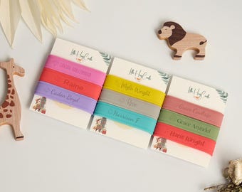 May include: Three sets of personalised silicone bracelets in pink, red, and purple, yellow, grey, and teal, and mauve, green, and red. Each bracelet has a name printed on it. A wooden giraffe and lion are also in the image.